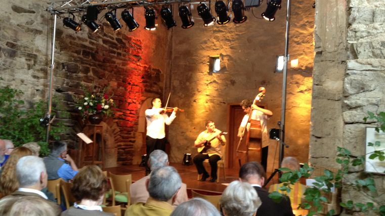 Ein musikalisches Konzert in einem alten Raum. Drei Musiker spielen auf der Bühne vor einem interessierten Publikum.