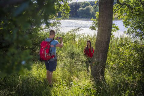 Zwei Wanderer stehen am Ufer eines Gewässers in einer grünen Umgebung. Der Mann mit einem roten Rucksack fotografiert die Frau, die in der Nähe eines Baumes steht.