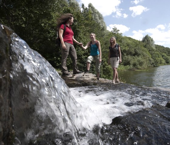 Drei Frauen wandern an einem kleinen Wasserfall in einem Waldsee entlang. Sie tragen Freizeitkleidung und Rucks&auml;cke. Der Himmel ist blau mit einigen Wolken., &copy; Traumpfade/Kappest