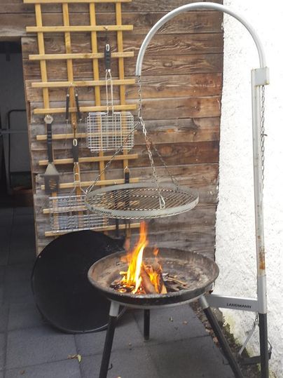 Ein Grill mit offenem Feuer steht auf einem gepflasterten Boden. Im Hintergrund ist eine Holzwand mit verschiedenen Grillutensilien zu sehen.