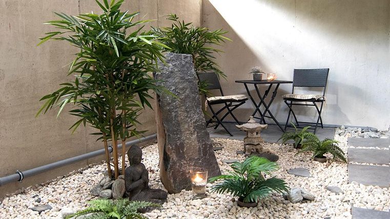 Ein ruhiger Innenhof mit grünem Bambus, Steinen und einer Buddha-Statue. Zwei Stühle und ein Tisch laden zum Entspannen ein.