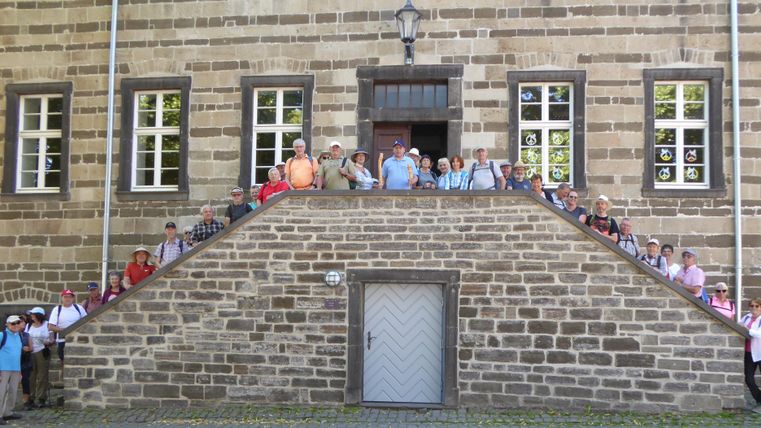 Eine Gruppe von Menschen steht auf einer Treppe vor einem historischen Gebäude. Der Sonnenlicht fällt auf die Fassade und die Teilnehmer haben fröhliche Gesichtsausdrücke.