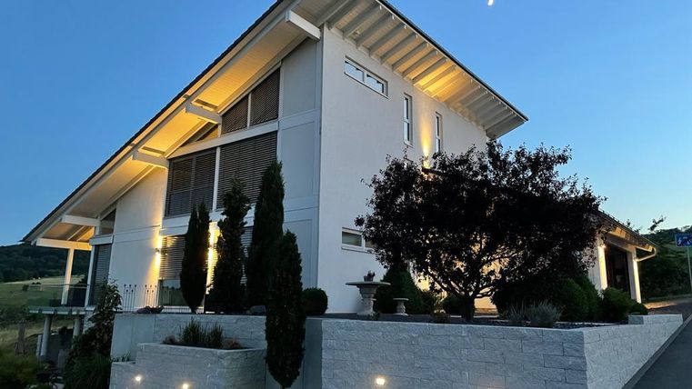 Ein modernes Haus mit einer eleganten Fassade und Außenbeleuchtung. Der Garten ist gepflegt und durch Bäume und Sträucher geschmückt.