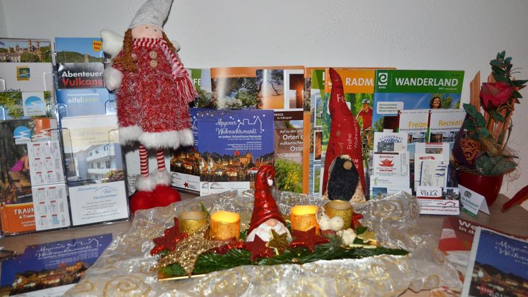 Eine festliche Dekoration mit Weihnachtsmännern und Kerzen steht auf einem Tisch. Im Hintergrund sind verschiedene Broschüren und Flyer aus einer Region zu sehen.