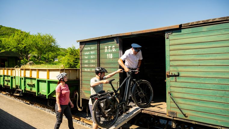 A man is carrying his bicycle into a green wagon while a woman watches. It is a sunny day with a blue sky and a lot of greenery in the background.