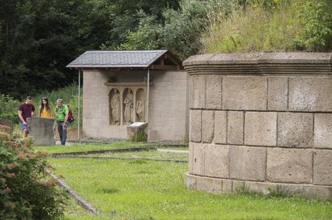 Eine Gruppe von Menschen spaziert in einer grünen Umgebung. Im Hintergrund sind steinerne Skulpturen und ein überdachter Bereich zu sehen.