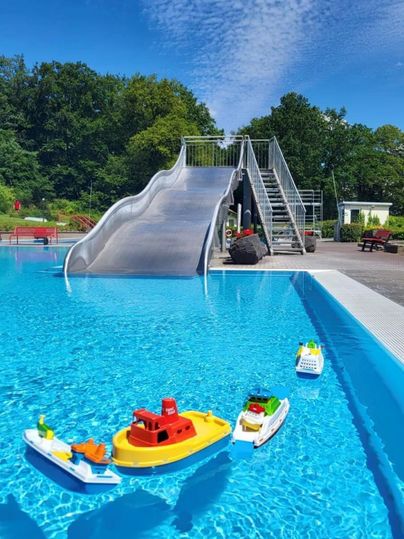 A swimming pool with a silver slide and colorful play boats. The sky is clear and the surroundings are green.