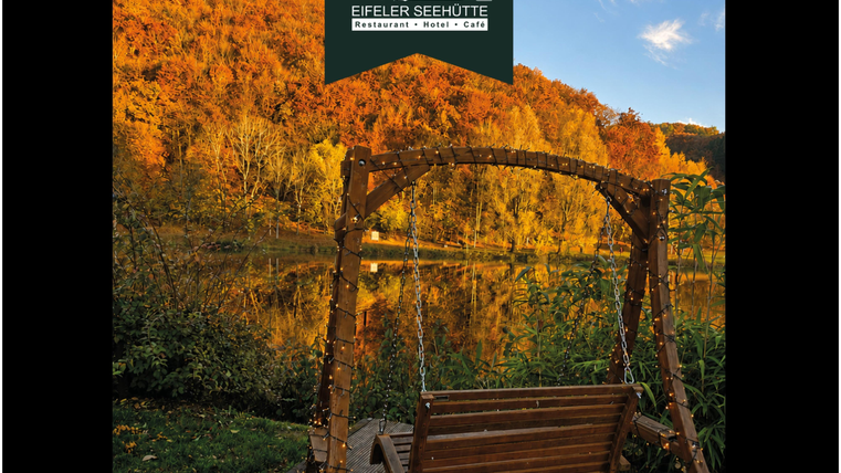Ein schaukelnder Holzstuhl steht am Ufer eines klaren Sees. Im Hintergrund leuchten die bunten Herbstblätter der Bäume.