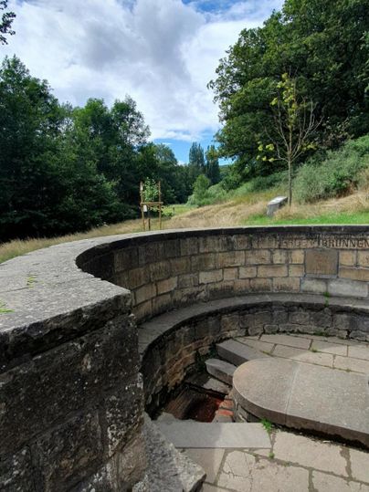 Eine steinerne Konstruktion mit runden Stufen in einer grünen Landschaft. Der Himmel ist teilweise bewölkt und es sind Bäume im Hintergrund zu sehen.