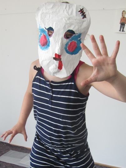Ein Kind trägt eine bunte Maske aus Papier und schaut direkt in die Kamera. Es steht in einem Raum und hebt die Hand auf eine spielerische Weise.