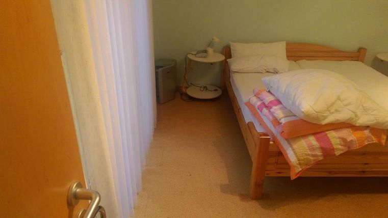 A simple bedroom with a wooden bed, pillows, and a small nightstand. The walls are in a soft color tone and there is a bright curtain.