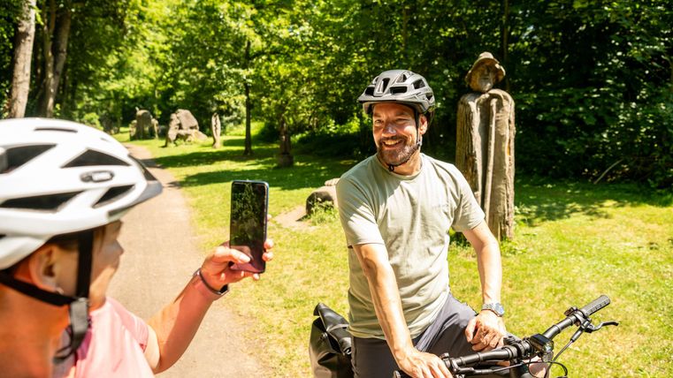 Zwei Radfahrer in einem grünen Park erfreuen sich an einem sonnigen Tag. Einer von ihnen macht ein Foto mit seinem Smartphone, während der andere lächelt.