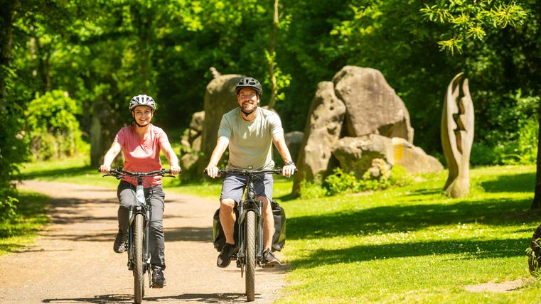 Zwei Personen fahren fröhlich mit dem Fahrrad auf einem Waldweg. Die Umgebung ist grün und sonnig, mit großen Steinskulpturen im Hintergrund.
