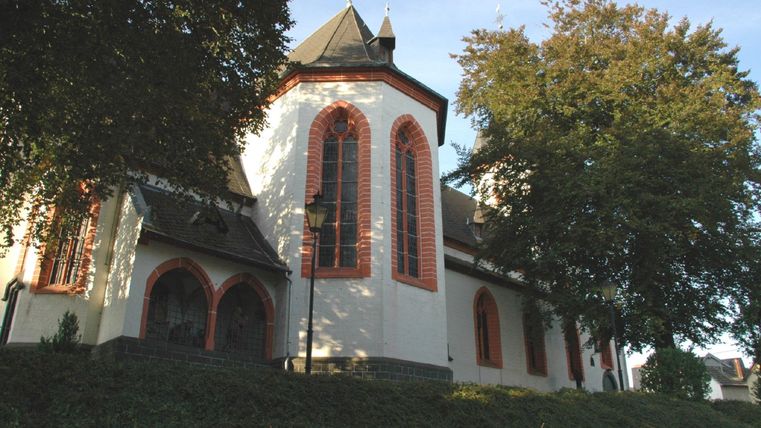 Eine hübsche Kirche mit roten Fenstern und einem schlichten Dach. Umgeben von Bäumen und einem grünen Hang.