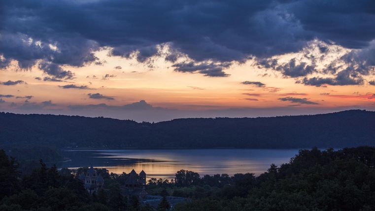 Ein schöner Sonnenuntergang über einem ruhigen See, umgeben von Hügeln. Der Himmel ist in orange und blau getönt, während Wolken vorbeiziehen.