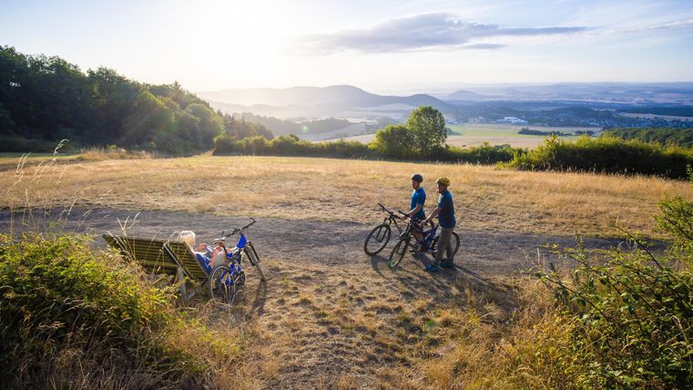 Zwei Radfahrer stehen auf einem Hügel und genießen die Aussicht. Die Landschaft ist weitläufig und von sanften Hügeln umgeben, während die Sonne aufgeht.
