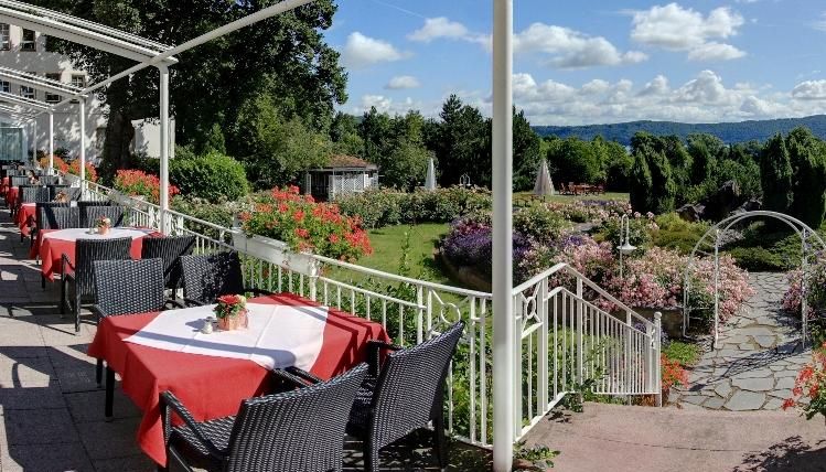 An inviting restaurant with red tablecloths and outdoor chairs. In the background, blooming gardens and a clear blue sky can be seen.