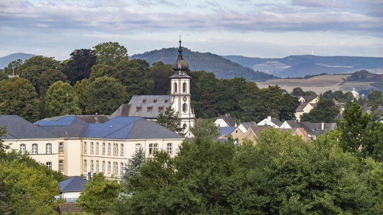 Eine ruhige Landschaft mit einem historischen Gebäude und einem Kirchturm. Im Hintergrund sind sanfte Hügel und Bäume zu sehen.
