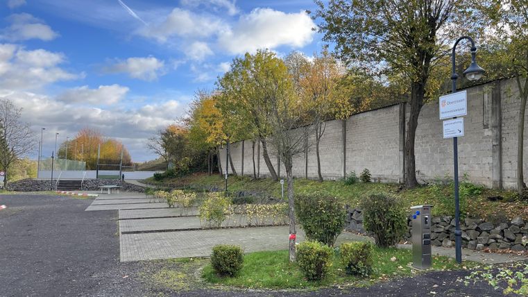 Ein ruhiger Park mit Bäumen und einer Mauer im Hintergrund. Der Himmel ist blau mit einigen Wolken.