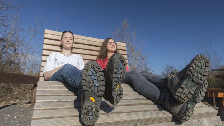 Two teenagers are sitting relaxed on a wooden lounge chair. They are wearing sturdy shoes and enjoying the sunny weather under a clear blue sky.