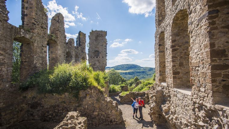 Eine alte Burgruine mit grünem Bewuchs und Aussicht auf die Landschaft. Zwei Personen stehen in der Ruine und genießen die Aussicht.