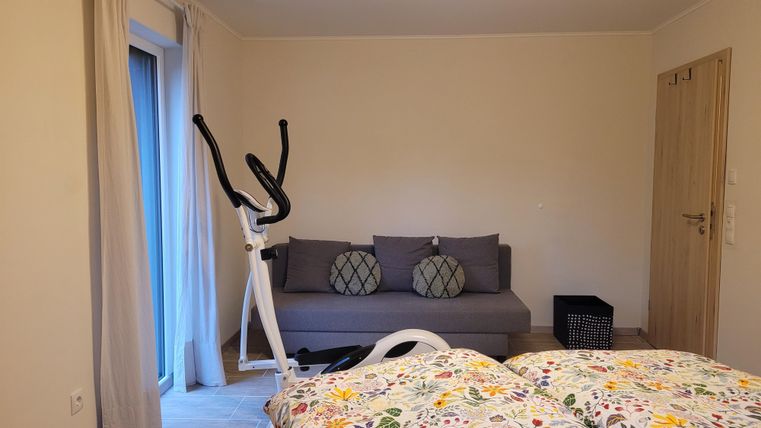 Ein gemütlicher Raum mit einem Sofa und einem Fitnessgerät. Es gibt helle Vorhänge und eine bunte Bettdecke auf einem Bett.