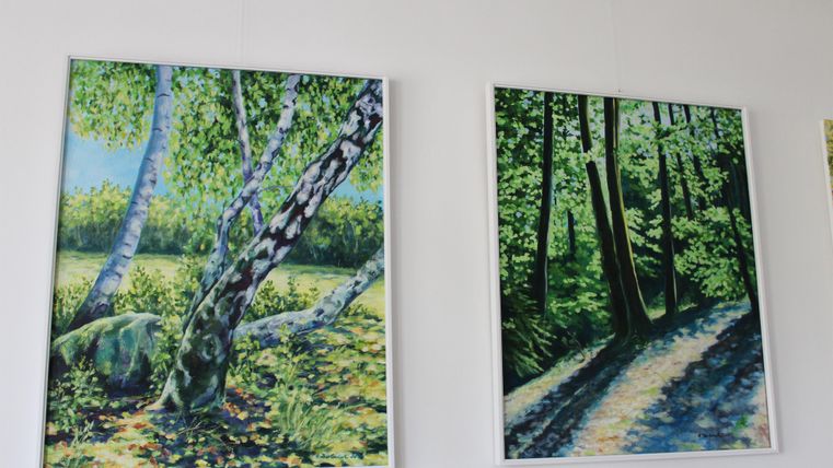 Zwei farbenfrohe Landschaftsbilder sind an der Wand. Das linke Bild zeigt birkenartige Bäume in einem sonnigen Umfeld, während das rechte Bild einen schattigen Weg durch einen Wald darstellt.