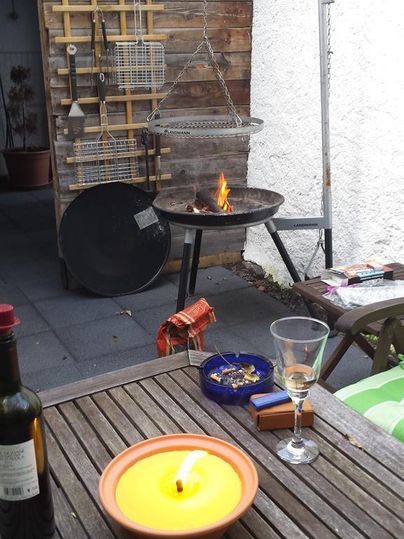 Ein gemütlicher Außenbereich mit einem Grill und flammendem Feuer. Auf dem Tisch stehen Wein, eine Kerze und ein Glas.