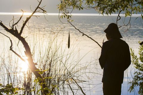 Ein Angler steht silhouettenhaft am Ufer eines ruhigen Sees. Die Sonne spiegelt sich auf der Wasseroberfläche und es umgibt ihn üppiges Grün.