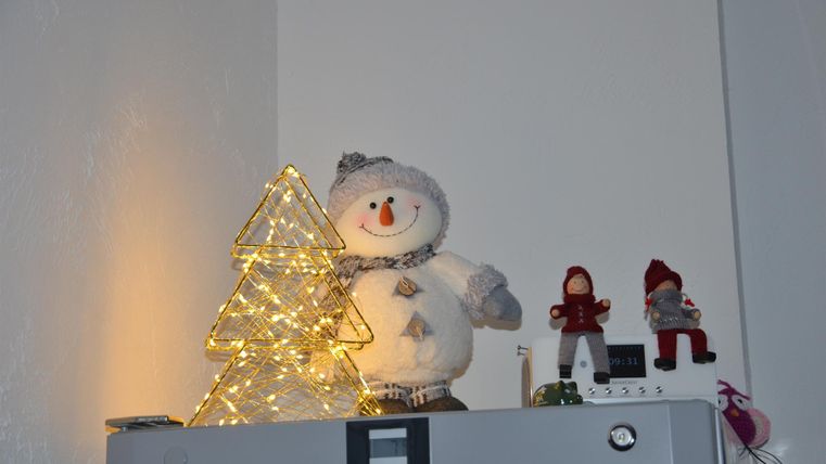 Ein dekorierter Kühlschrank mit einem leuchtenden, dreidimensionalen Weihnachtsbaum und einem niedlichen Schneemann. Daneben stehen zwei kleine Figuren in festlicher Kleidung.