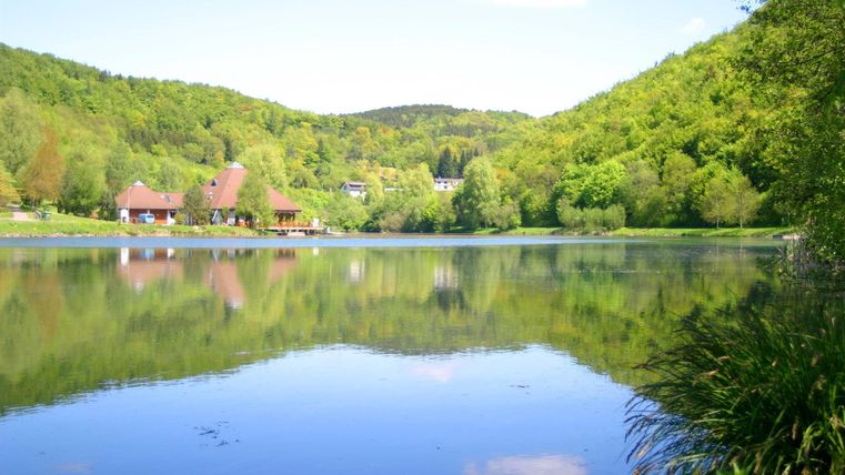 Ein ruhiger See umgeben von grünen Hügeln und Bäumen. Im Hintergrund sind einige Gebäude zu sehen.
