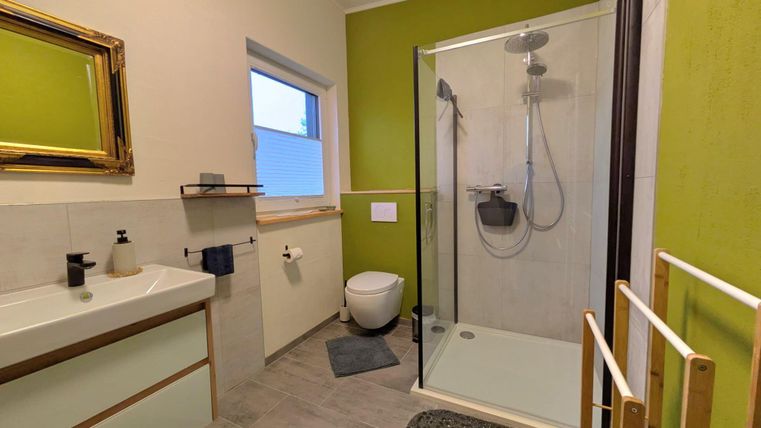 Ein modernes Badezimmer mit einer Glasdusche und grünen Wänden. Es gibt einen Waschtisch, ein WC und große Fenster für natürliches Licht.