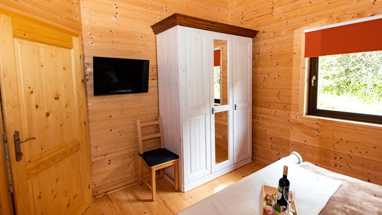 A cozy wooden room with a wardrobe, a television, and a bed. The window offers a view of nature.