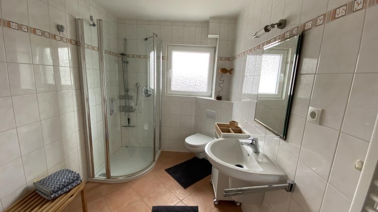 Ein modernes Badezimmer mit einer Duschkabine und einem Waschbecken. Das Design ist hell und freundlich mit weißen Fliesen und einem Holzstuhl.