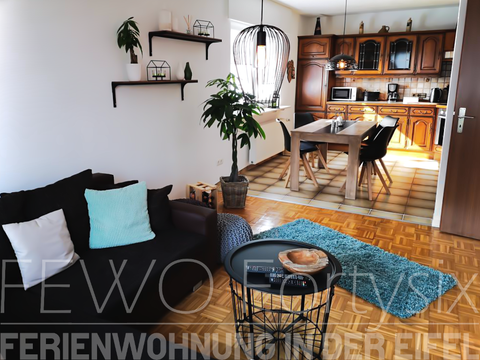 Ein gemütliches Wohnzimmer mit einer schwarzen Couch und einem stilvollen Tisch. Im Hintergrund befindet sich eine moderne Küche und ein Essbereich mit Holzstühlen.
