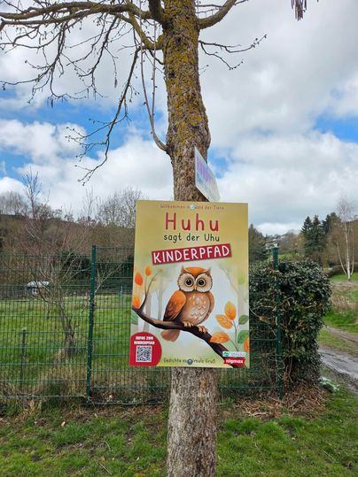 A sign on a tree with the inscription "Hoo says the owl children's path." The sign features an illustration of an owl with colorful flowers.
