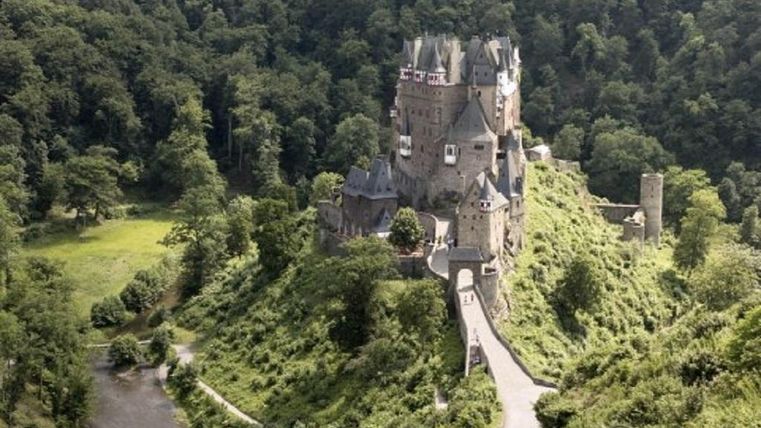 Eine beeindruckende Burg auf einem Hügel, umgeben von grünen Wäldern. Ein kleiner Fluss fließt in der Nähe der Burg.