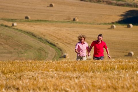 Ein Paar läuft Hand in Hand durch ein goldenes Feld mit Heuballen im Hintergrund.