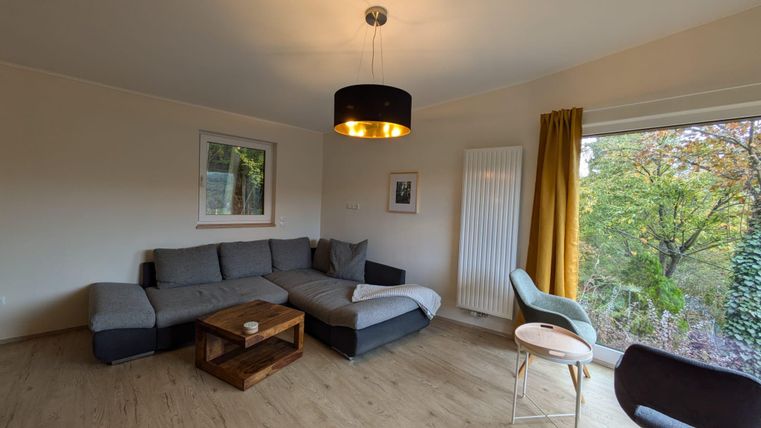 Ein gemütliches Wohnzimmer mit einem großen grauen Sofa und einem Holztisch. Durch ein Fenster ist eine grüne Landschaft zu sehen.