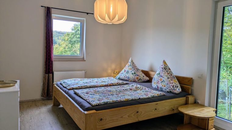Ein helles Schlafzimmer mit einem großen Bett und bunten Bettwäsche. Durch das Fenster ist viel Tageslicht und Blick ins Grüne.