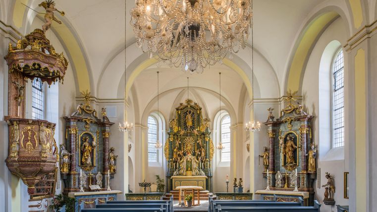 A beautiful church with high ceilings and an impressive chandelier. The benches are neatly arranged in rows, and the walls are adorned with intricate statues.