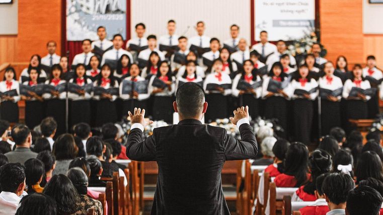 A choir sings in a church, led by a conductor. In the background, many listeners can be seen attentively listening to the performance.