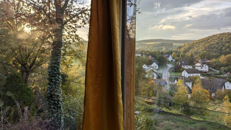 Ein Blick aus dem Fenster auf eine malerische Landschaft mit Bäumen und sanften Hügeln. Im Vordergrund ist ein gelbes Vorhangteil sichtbar.