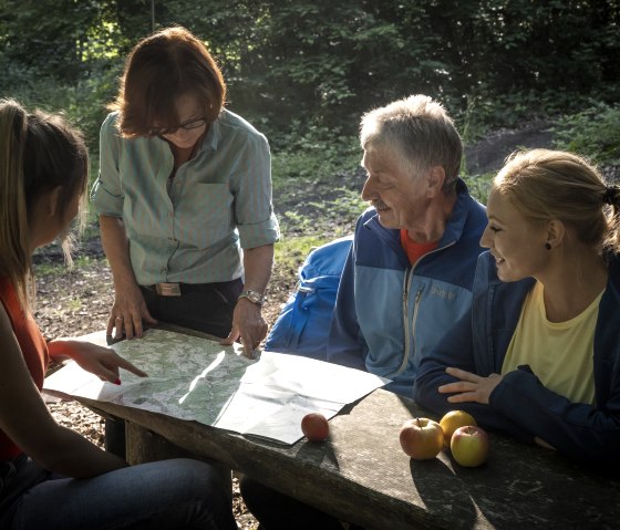 Four people are looking at a map at a wooden table in the forest. There are apples on the table. Sunlight falls through the trees., © Kappest_REMET