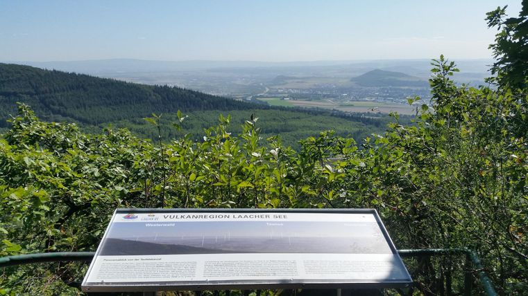 Eine Aussicht auf eine grüne Landschaft mit sanften Hügeln und einem klaren Himmel. Im Vordergrund steht ein Informationsschild über den Vulkansee.