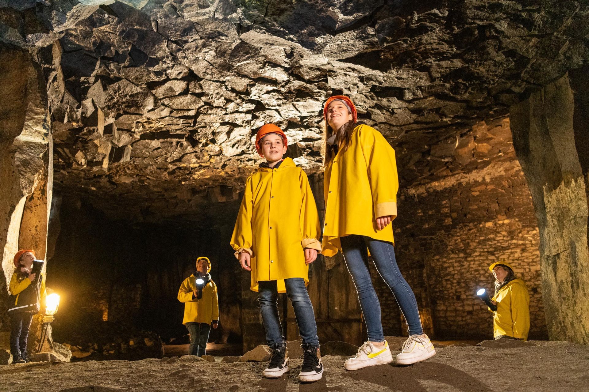 Two children in yellow rain jackets are standing in a cave. In the background, more people with flashlights can be seen.