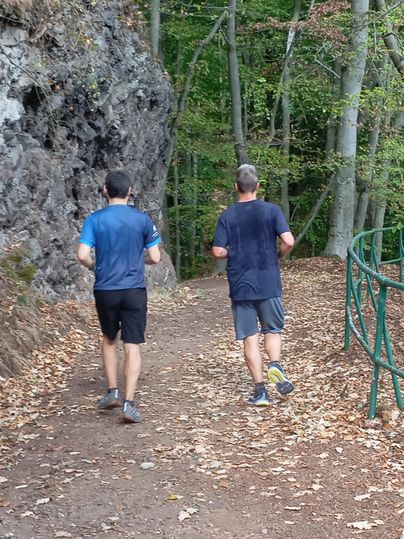 Twee mannen joggen op een bospad naast een rotswand. De omgeving is gekenmerkt door bomen en herfstbladeren.