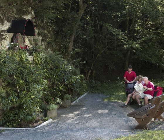 Drei Personen sitzen auf einer Bank im Wald, betrachten eine Karte. Links ist eine Madonna-Statue in einer Felswand zu sehen, umgeben von Pflanzen., © Traumpfade/Kappest