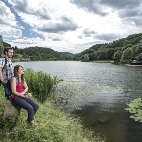 Two people are sitting on the shore of a lake, surrounded by green hills and trees. A house stands on the right bank. The sky is cloudy., © Kappest/REMET