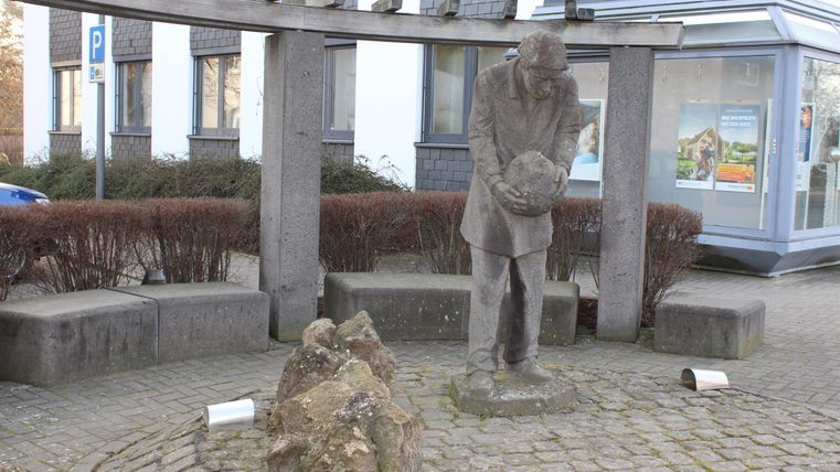 A statue of a man holding a stone. In the background, modern buildings and shrubs are visible.